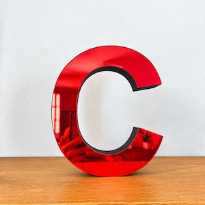 Può includere: Una lettera C tridimensionale con una superficie frontale rossa lucida e riflettente e un bordo laterale nero solido. La lettera ha una finitura liscia e lucida che crea un effetto specchio, mostrando sottili riflessi dell'ambiente circostante.