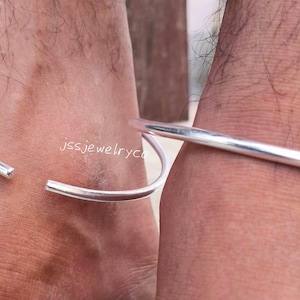 以下が含まれることがあります： 足首を囲む、オープンデザインのシルバーアンクレット2つ。アンクレットは滑らかで光沢のある金属製です。画像は人の下肢と足を示しています。