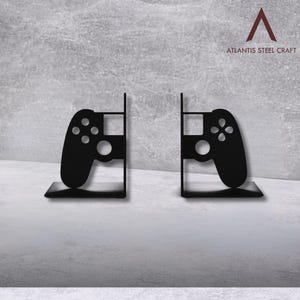 May include: Black metal bookends shaped like video game controllers. Each bookend features a controller silhouette with buttons and a central circular design. The text "ATLANTIS STEEL CRAFT" is visible in the upper right corner.