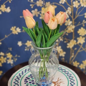 May include: A clear glass vase with a fluted design holds a bouquet of tulips. The tulips have peach and pink petals and green stems. The vase sits on a round table with a decorative placemat, against a blue floral wallpaper background.