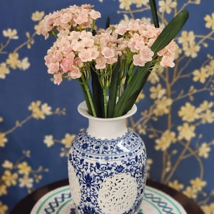 May include: A white and blue ceramic vase with a floral design, filled with pink flowers and green leaves. The vase has a detailed pattern and is placed on a round table with a decorative mat. The background is a blue wall with a floral pattern.