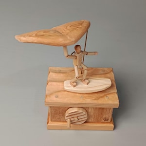 May include: A light-colored wooden mechanical automaton featuring a small articulated figure standing on a surfboard. The figure holds a large, curved wooden wing above its head. The base is a rectangular wooden box with a circular hand crank on the side.