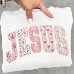 May include: White sweatshirt with the word "JESUS" appliqued across the front. The letters are made of pink floral fabric with a scalloped edge. The sweatshirt is a crew neck style.