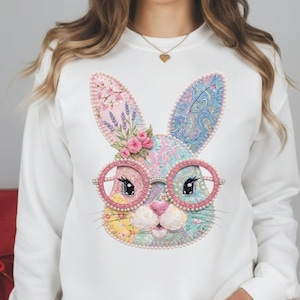 May include: White sweatshirt featuring a colorful patchwork bunny face with pink-rimmed glasses. The bunny has floral accents and a pink nose. The ears are decorated with lavender and paisley patterns. A whimsical design for Easter or spring.