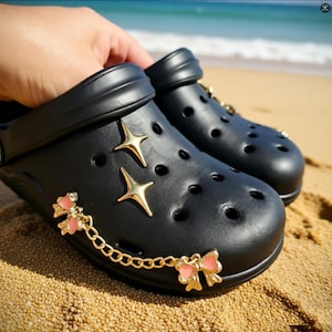 Peut inclure: Sabots noirs ornés d'étoiles dorées et de breloques en forme de nœuds roses. Les chaussures sont décorées d'étoiles dorées et de nœuds roses reliés par une chaîne dorée. Les sabots sont posés sur du sable.