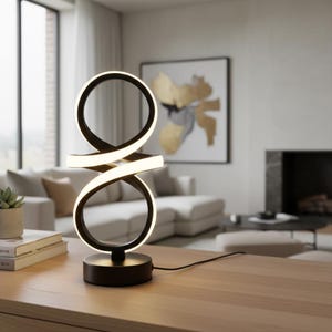 Peut inclure: Lampe de table LED noire et blanche moderne avec un design unique en forme de boucle infinie. La lampe a une base circulaire et une forme de ruban continu et éclairé. La lumière est chaleureuse, idéale pour un salon ou une chambre. La lampe mesure environ 30 cm de haut.