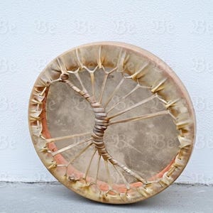May include: A circular drum with a natural hide head and a woven handle. The drum has a light brown frame, and the hide is a lighter tan. The drum is likely used for music or spiritual practices, with a diameter of approximately 30 cm.