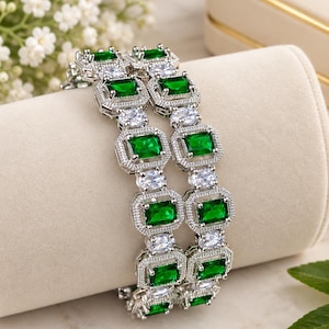 May include: Two silver-toned bangles featuring a repeating pattern of rectangular emerald-green stones alternating with smaller, clear oval-cut stones. Each green stone is framed by a border of tiny, clear pavé-set stones, creating a structured, linked design.
