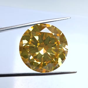 Op de afbeelding: Een grote, ronde, gefacetteerde gele edelsteen wordt door een pincet tegen een witte achtergrond gehouden. De edelsteen heeft een briljante slijpvorm, die licht reflecteert en een sprankelend effect creëert. De kleur is een warm, goudgeel.