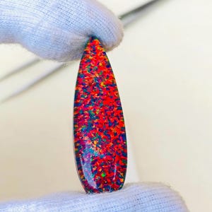 May include: A teardrop-shaped opal gemstone featuring a vibrant mosaic of red, blue, green, and orange flecks. The polished stone displays a high-contrast play of color across its surface, set against a dark base, showcasing a smooth, reflective finish.