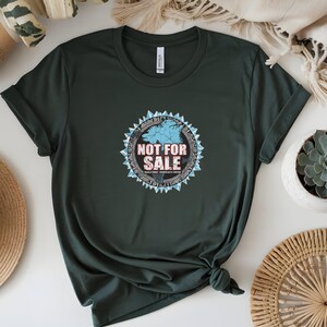 May include: Dark green t-shirt featuring a circular graphic with the text "NOT FOR SALE". The graphic includes a blue map design, surrounded by a white and blue geometric border. The shirt is knotted at the bottom.