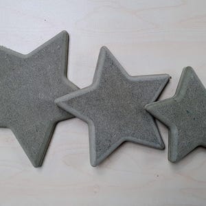 May include: Three grey, star-shaped decorative objects of varying sizes. The stars are made of a textured material and are arranged on a light-coloured surface. The largest star is approximately 25 cm across.