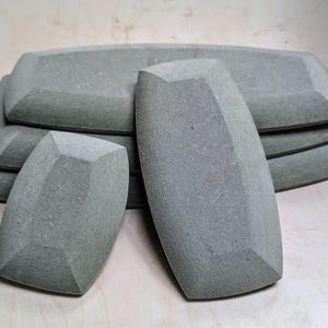 Może przedstawiać: Zestaw szarych, kamiennych półmisków i mydelniczek. Półmiski są prostokątne ze ściętymi krawędziami, a mydelniczki owalne z fasetowanymi bokami. Przedmioty ułożone są na jasnej drewnianej powierzchni.