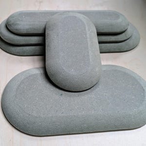 May include: A set of grey, oval-shaped stone coasters. The coasters are arranged in a tiered stack, with one standing upright. The stone has a textured surface and rounded edges. These coasters are designed for drinks and beverages.