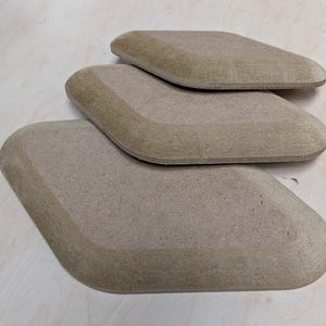 May include: Three diamond-shaped wooden objects in shades of light brown are stacked. The edges of each piece are a slightly darker tone than the centre, creating a subtle border. The objects are arranged on a light-coloured wooden surface.