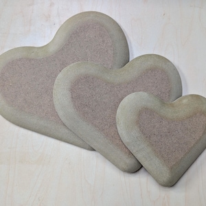 Może przedstawiać: Trzy dekoracyjne przedmioty w kształcie serca o różnych rozmiarach. Każde serce ma jasnozieloną obwódkę i teksturowane, stonowane różowe wnętrze. Serca ułożone są na jasnej drewnianej powierzchni, tworząc efekt warstwowy.