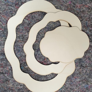 May include: A selection of unfinished wooden cutouts in diverse organic forms. The set comprises two wavy circular frames and two cloud-shaped pieces, all with a natural wood grain. They are arranged on a textured, multi-coloured fabric.