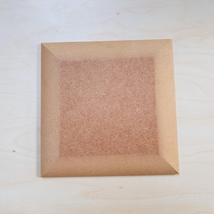 Może przedstawiać: Kwadratowa, niewykończona drewniana tablica ze ściętą krawędzią. Tablica jest w jasnobrązowym kolorze i wydaje się być wykonana z płyty wiórowej lub podobnego materiału. Krawędzie są nieco ciemniejsze niż środek.