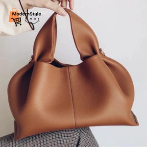 May include: A brown leather handbag with a rounded shape and two handles. The bag has a textured surface and a vertical seam down the center. The bag is held up by a hand.