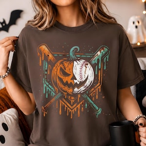 May include: A dark brown t-shirt featuring a graphic print of a split pumpkin and baseball design. The central image shows a pumpkin on the left and a baseball on the right, crossed by two baseball bats with a dripping, distressed paint effect.