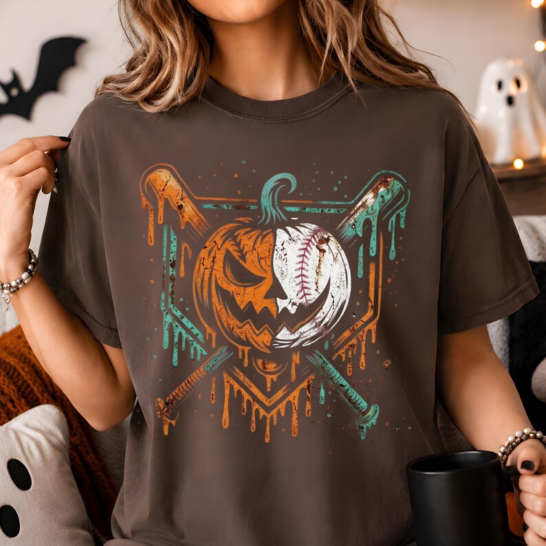 May include: A dark brown t-shirt featuring a graphic print of a split pumpkin and baseball design. The central image shows a pumpkin on the left and a baseball on the right, crossed by two baseball bats with a dripping, distressed paint effect.