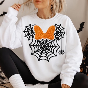 May include: A white long-sleeve sweatshirt featuring a graphic of three black spiderwebs arranged in the shape of a mouse head. A glittery orange bow is centered at the top, and a small black spider hangs from one of the webs on the right side.