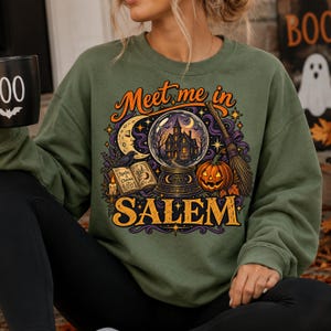 May include: An olive green crewneck sweatshirt featuring a graphic print with the text "Meet me in SALEM". The design includes a crystal ball with a house inside, a crescent moon, a spellbook, a candle, a broomstick, and a jack-o'-lantern.