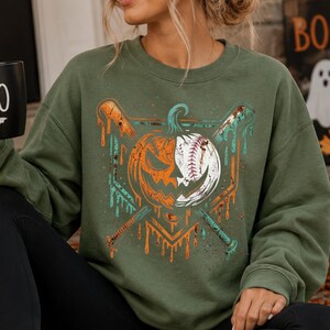 May include: An olive green crewneck sweatshirt featuring a graphic print of a split pumpkin and baseball design. The graphic includes two crossed baseball bats with a dripping paint effect in orange and teal colors, set against the solid fabric.