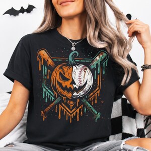 May include: A black t-shirt featuring a graphic print of a split pumpkin and baseball design. The pumpkin is orange with a carved face, while the other half is a white baseball. Two crossed baseball bats are positioned behind the central image.