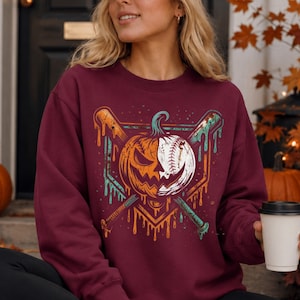 May include: A maroon crewneck sweatshirt featuring a graphic print of a split pumpkin and baseball design. The central image shows a jack-o'-lantern on one side and a baseball on the other, crossed by two baseball bats with a dripping paint effect.