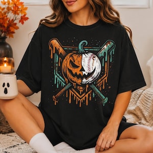 May include: A black t-shirt featuring a graphic print of a pumpkin and baseball hybrid design split down the middle, positioned over two crossed baseball bats. The artwork includes dripping paint effects in orange and teal colors on a dark background.
