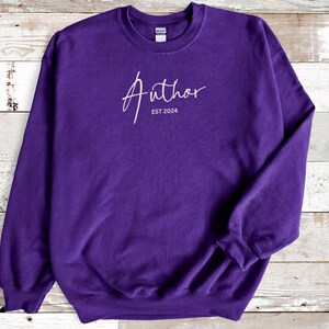 May include: A purple sweatshirt with the word "Author" in a cursive font, and the text "EST 2024" below it. The sweatshirt has a crew neck and long sleeves.