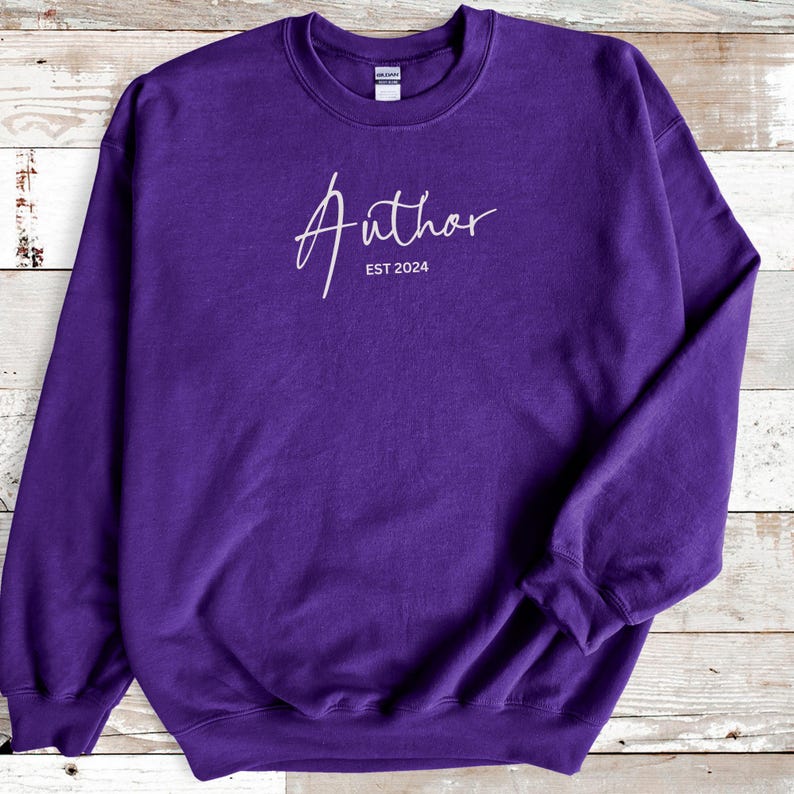 May include: A purple sweatshirt with the word "Author" in a cursive font, and the text "EST 2024" below it. The sweatshirt has a crew neck and long sleeves.