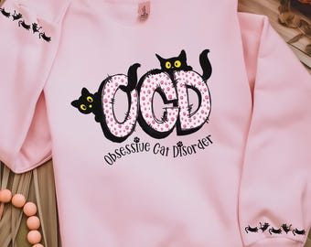 May include: A light pink sweatshirt with the text "OCD Obsessive Cat Disorder". The letters "OCD" are in black with a pink paw print pattern. Black cat silhouettes are on the sleeves.
