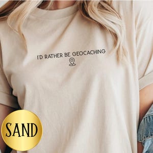 May include: Sand-colored t-shirt with the text "I'D RATHER BE GEOCACHING" and a location pin graphic. The shirt is a neutral color and has a casual, relaxed fit. The word "SAND" is printed on a gold circle.