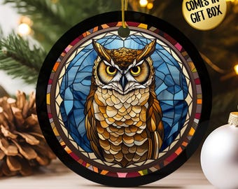 May include: Round Christmas ornament featuring a detailed owl design. The owl has brown and tan feathers, yellow eyes, and is set against a blue stained-glass background. The ornament has a black border and a gold hanging string. The text "COMES IN GIFT BOX" is on the top right.