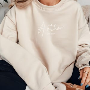 May include: Beige sweatshirt with the word "Author" in white script, with "EST 2024" below it. The sweatshirt has a crew neck and long sleeves. The person wearing the sweatshirt is also wearing a ring on their finger.