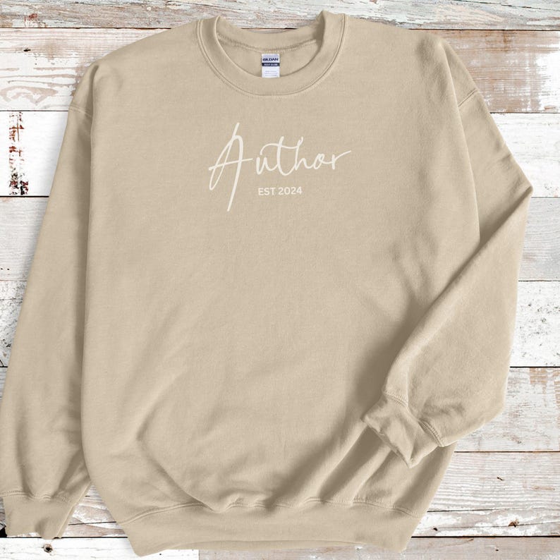 May include: A tan-colored sweatshirt with the word "Author" in a cursive font, and the text "EST 2024" below it. The sweatshirt has a crew neck and long sleeves.