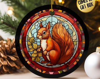 May include: A round Christmas ornament featuring a stained-glass design with a squirrel. The squirrel is depicted in shades of brown and orange, holding leaves. The ornament has a black border and a gold string for hanging. Text on the image says "COMES IN GIFT BOX".