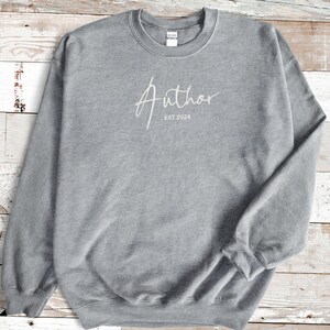 May include: A gray sweatshirt with the word "Author" in a cursive font and the text "EST 2024" printed in white. The sweatshirt has a crew neck and long sleeves. The background is a light-colored wooden surface.