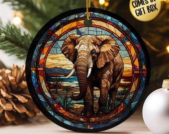 May include: Round Christmas ornament featuring a detailed illustration of an elephant in a stained-glass style. The ornament has a black border and a gold hanging string. The background depicts a landscape with water and a sunset. The text "COMES IN GIFT BOX" is on the top right.