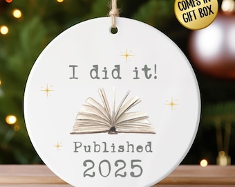Op de afbeelding: Ronde witte keramische ornament met de tekst "I did it! Published 2025" en een illustratie van een open boek. Het ornament heeft een touwtje om op te hangen en is versierd met gouden sterren. Een gele cirkel met de tekst "COMES IN GIFT BOX" is zichtbaar.