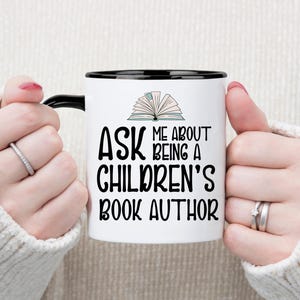 May include: White coffee mug with a black rim and handle. The mug has a black and white illustration of an open book and the text "Ask me about being a children's book author."