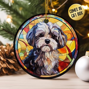 May include: Round Christmas ornament featuring a dog portrait in a stained-glass style. The dog has brown, black, and white fur, set against a colorful background. The ornament has a black border and a gold string for hanging. The text "COMES IN GIFT BOX" is in a gold circle.