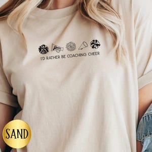 May include: Sand-colored t-shirt with the text "I'D RATHER BE COACHING CHEER" in black. The design includes cheerleading-related graphics. The shirt is made of a soft material.