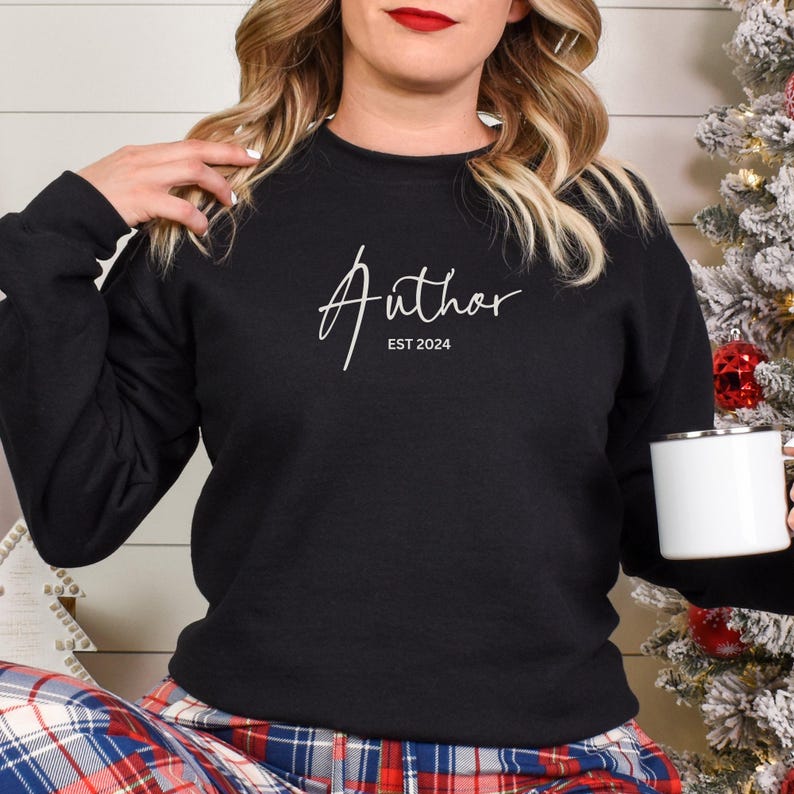 May include: Black sweatshirt with the word "Author" in white script and "EST 2024" below it. The person is holding a white mug. The person is wearing plaid pajama pants.
