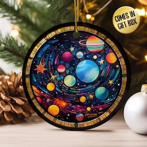 May include: Round Christmas ornament with a colorful galaxy design. The ornament features planets, stars, and a swirling nebula in shades of blue, purple, and orange. The text "COMES IN GIFT BOX" is on the upper right.