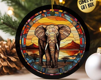 May include: Round black Christmas ornament featuring a stained-glass style elephant design. The elephant is brown with white tusks, set against a sunset landscape. The ornament has a gold string for hanging. Text on the ornament says "COMES IN GIFT BOX".
