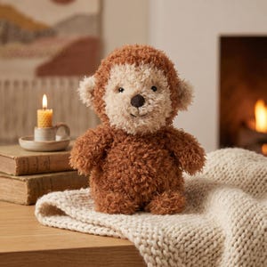 Peut inclure: Une peluche singe avec une fourrure marron et un visage, des oreilles et une poitrine cr&egrave;me. Le singe en peluche est assis sur une couverture en tricot cr&egrave;me. Une bougie allum&eacute;e et des livres sont en arri&egrave;re-plan.