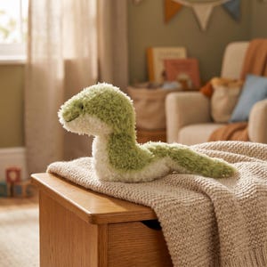 May include: A plush green and white snake toy rests on a beige blanket atop a wooden chest. The snake has a soft, textured appearance with a smiling face. The background includes a window, a chair, and decorative items, suggesting a nursery or playroom setting.